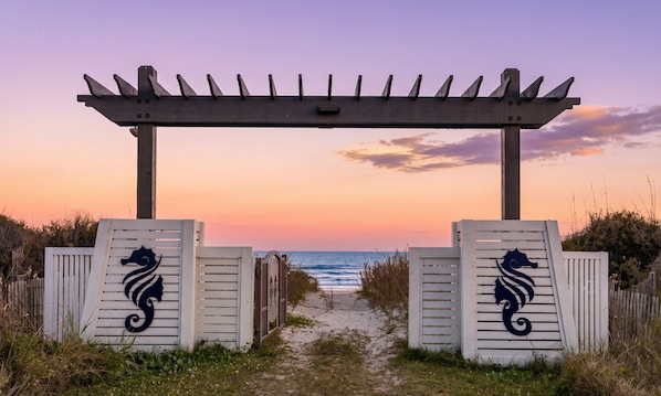 A wooden pergola entrance with a seahorse design, leading to a beach path.