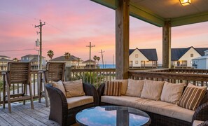 A wooden deck with wicker furniture and a glass table, overlooking a beach with houses and a sunset.