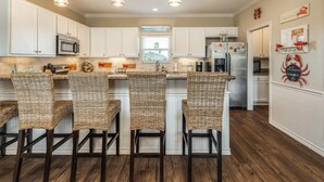 A kitchen with white cabinets, a stainless steel refrigerator, and a microwave. There are wicker chairs and a window with blinds.