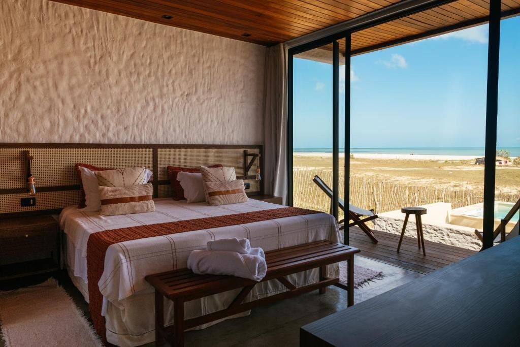 Quarto com uma cama grande, um banco de madeira e vista para a praia.