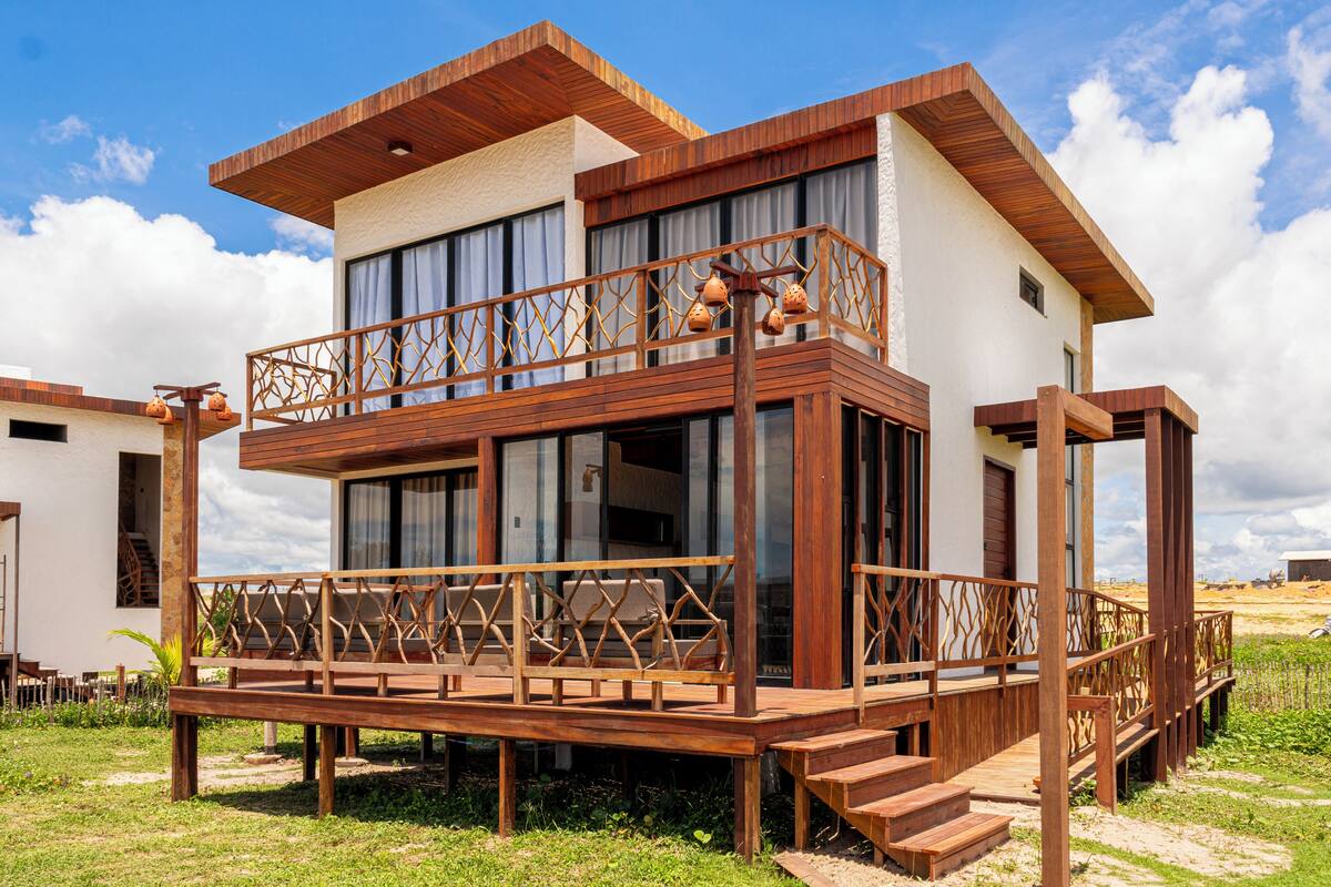 Un edificio moderno de dos pisos con terraza de madera, ventanas grandes y un balcón con detalles decorativos.