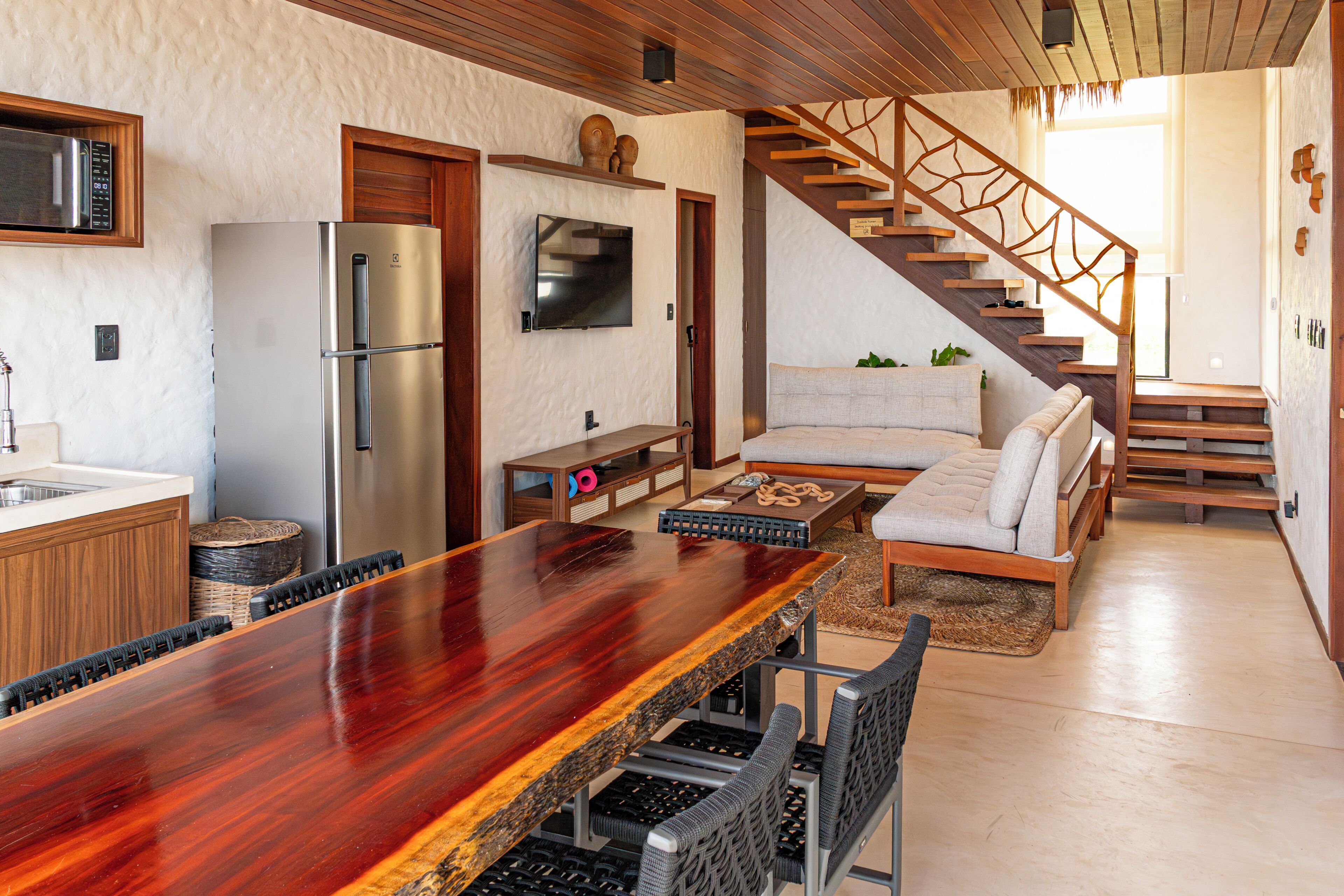 A modern kitchen with a wooden dining table, a built-in refrigerator, and a staircase leading to a living area with a sofa.