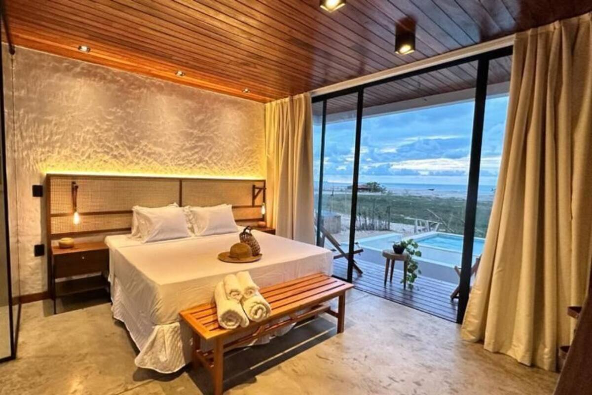 Un dormitorio con una cama grande, cabecera de madera, mesitas de noche, un banco y vistas al exterior.