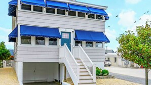 A two-story building with a blue awning, a white front door, and a staircase leading to the entrance. - Normandy Beach Retreat! Cozy 3BR Apartment, Just Steps from the sand (Lavallette)