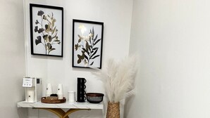 Un petit bureau à cadre blanc, doté d’un socle en fibres tressées, une chaise blanche et un vase orné de plumes blanches. Deux œuvres d’art florales encadrées sont accrochées au mur au-dessus du bureau.
