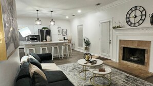 Un salon moderne avec un canapé noir, une cheminée, un coin repas et une cuisine en arrière-plan.