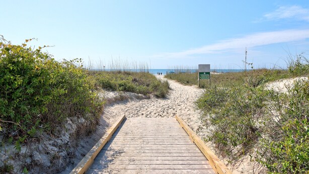 A wooden boardwalk leading to a beach with sand dunes and sparse vegetation.