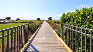 A wooden boardwalk with metal railings, surrounded by greenery and leading to a distant view.