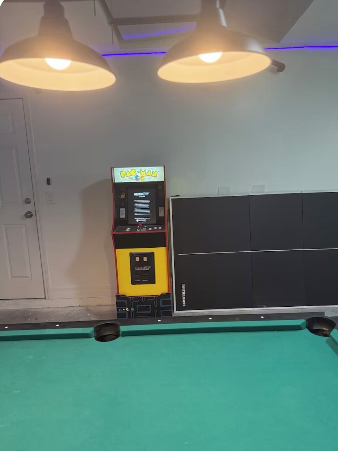 A pool table with overhead lighting, a Pac-Man arcade machine, and a black cabinet.