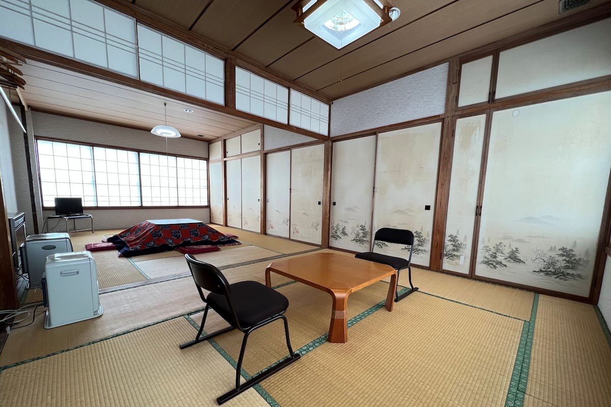 A traditional Japanese room with tatami flooring, a low wooden table, two black chairs, a TV, and sliding doors with artwork.