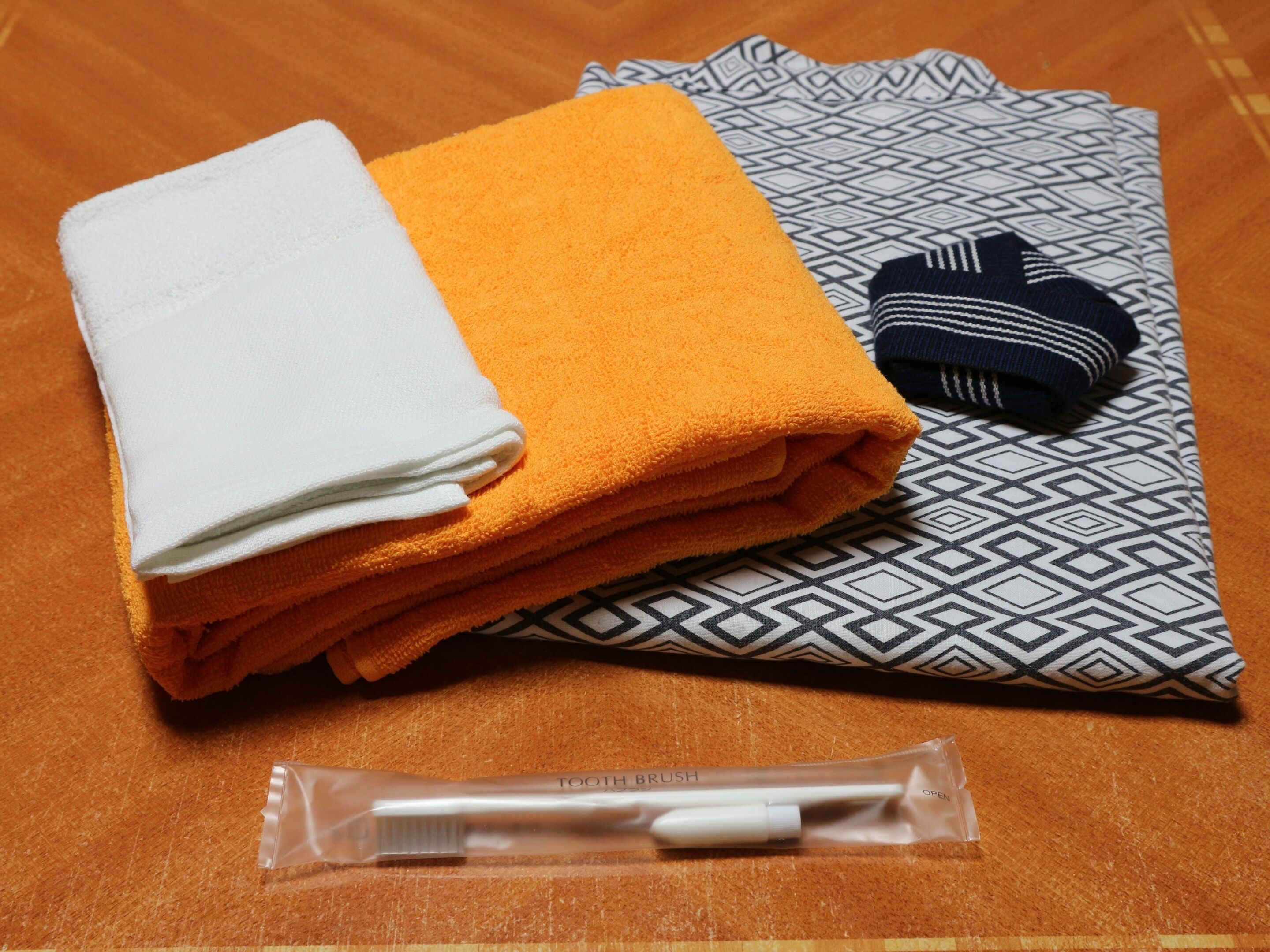 A neatly folded white and orange towel, a patterned pillow, a dark blue and white striped item, and a toothbrush in its packaging.