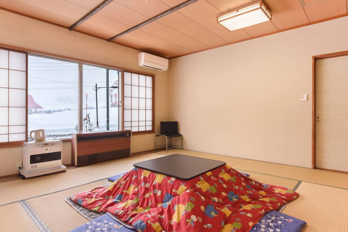 A traditional Japanese room with tatami flooring, a tatami mat table, a heater, and a view of a snowy landscape through large windows.