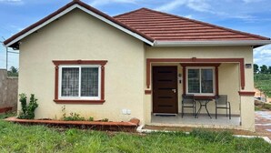 Uma casa térrea com telhado marrom, paredes bege e varanda frontal com duas cadeiras.