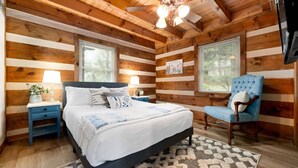 A wooden cabin bedroom with a bed, two nightstands, a blue chair, and a patterned rug.