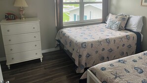 Quarto com cama, cômoda, janela com cortinas e vista para uma casa e vegetação lá fora.