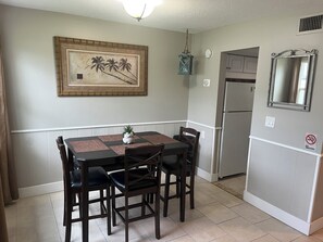 A dining area with a table, chairs, a painting, a hanging lantern, and a refrigerator.
