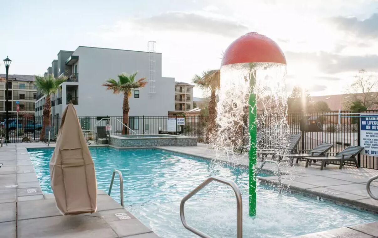 A swimming pool with a water feature, surrounded by palm trees and modern buildings.