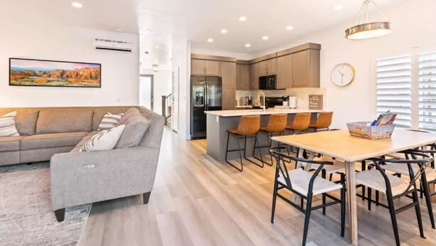 A modern living room with a grey sofa, a dining area with a wooden table and chairs, and a kitchen with a refrigerator and microwave.