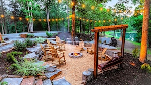 Un espace extérieur aménagé avec des chaises en bois et un foyer, entouré d’arbres et bordé d’un chemin de gravier.