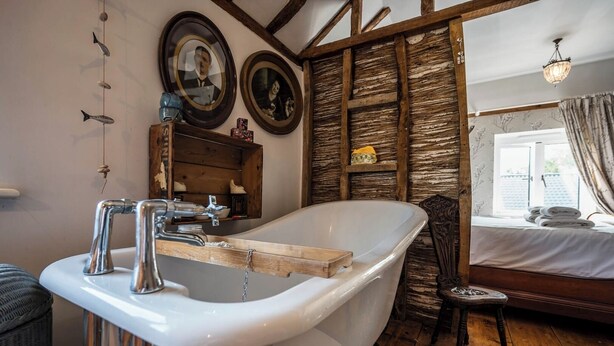 A bathroom with a freestanding bathtub, wooden beams, and a window with curtains. - Farm View suite, Kelsale (Kelsale)