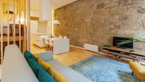 A modern living room with a stone wall, a sofa with cushions, a TV, and a dining area with a white table and chairs.