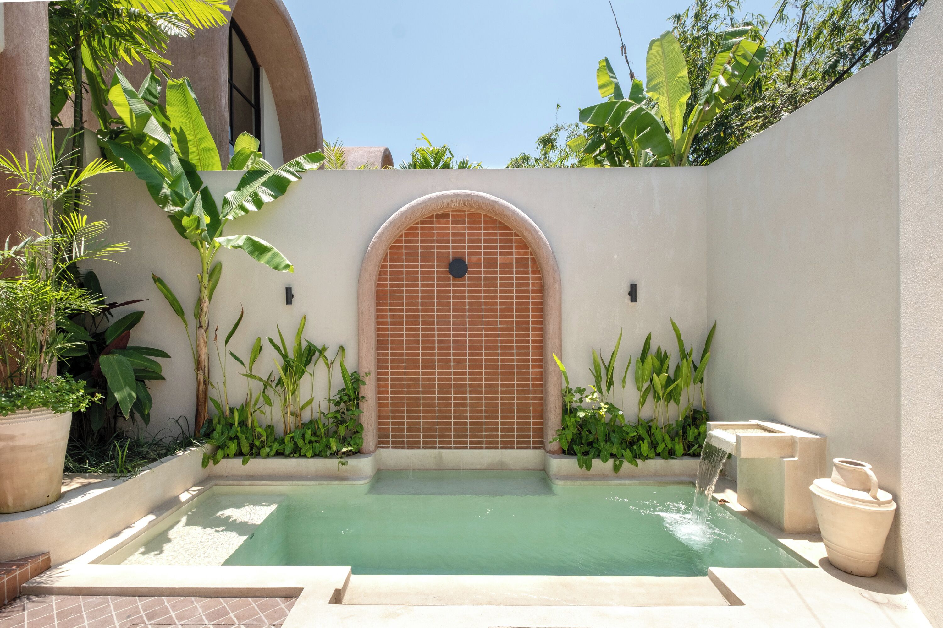 A small, serene pool area with a tiled wall, potted plants, and a stone bench.
