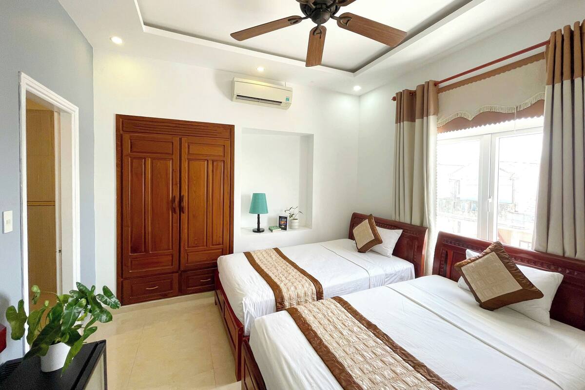 Habitación de hotel con dos camas, ventilador de techo, aire acondicionado, ventana con cortinas y una planta en maceta.