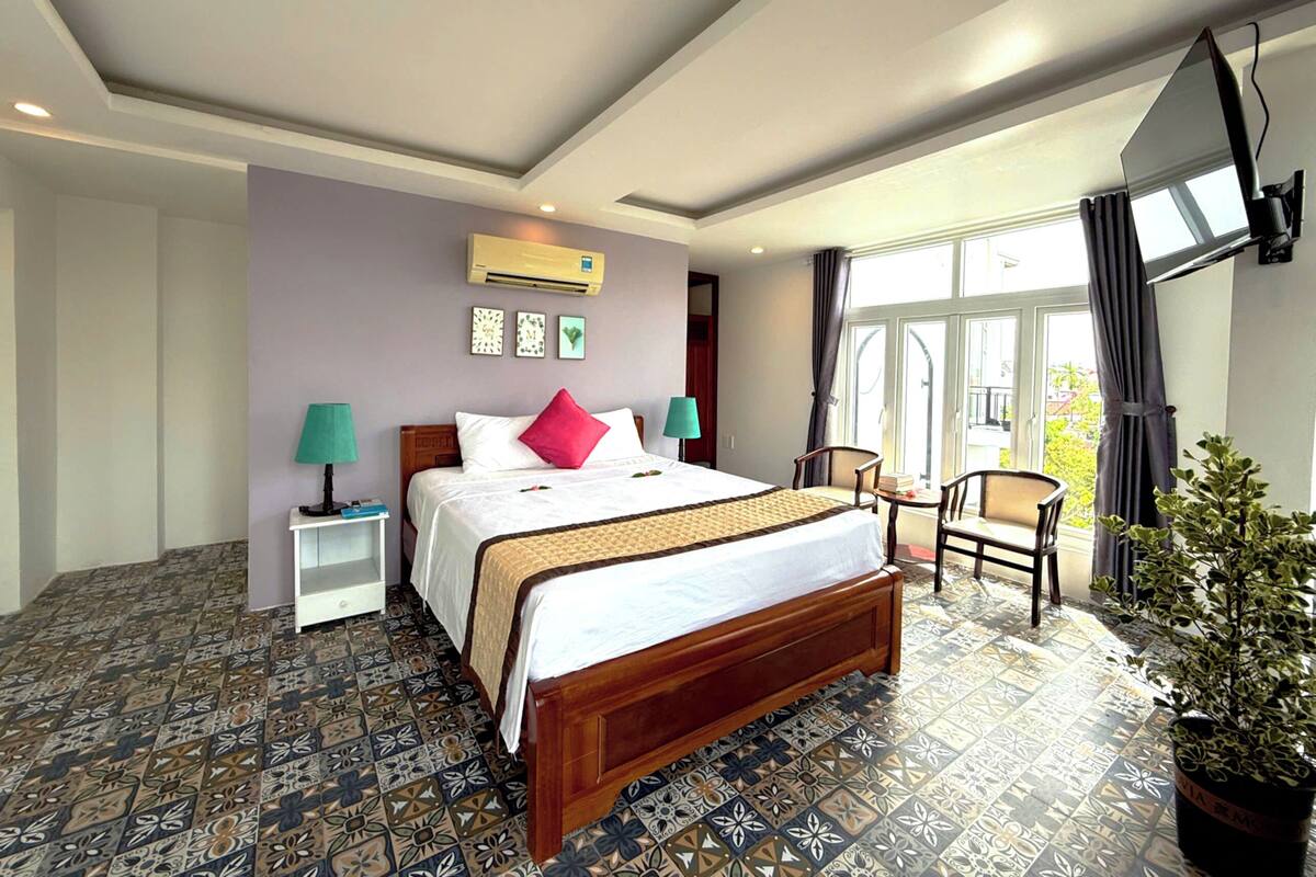 Habitación de hotel con una cama grande, dos mesitas de noche con lámparas, una pequeña mesa con sillas, una planta en maceta y un ventanal con cortinas.