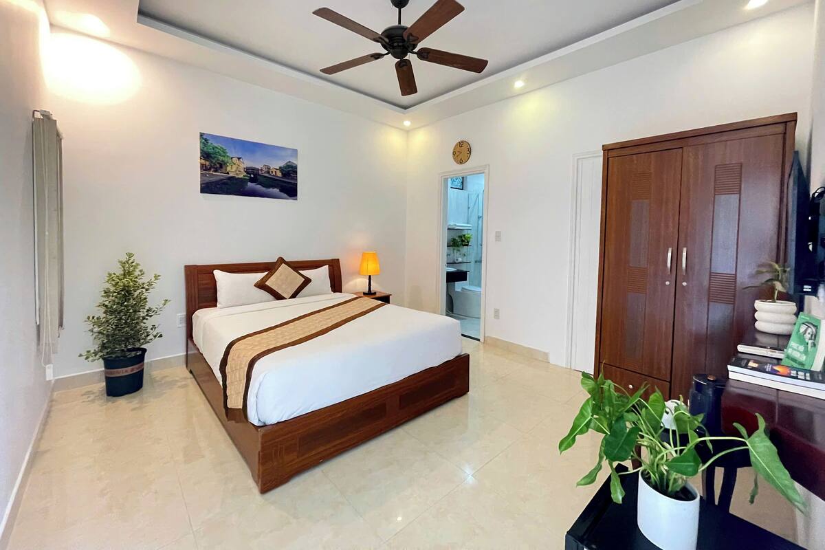 Habitación de hotel con una cama, ventilador de techo, un reloj, un cuadro, una planta en maceta y un armario.