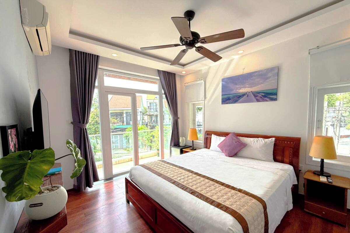 Habitación de hotel con cama, ventilador de techo, ventana grande con cortinas, mesita de noche con lámpara y una planta.