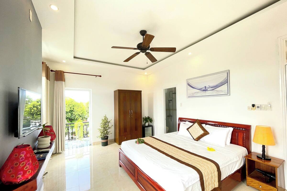 Habitación de hotel con cama, mesita de noche, lámpara, ventilador de techo, televisor y vista al exterior.