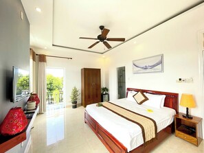 Habitación de hotel con cama, mesita de noche, lámpara, ventilador de techo, televisor y vista al exterior.