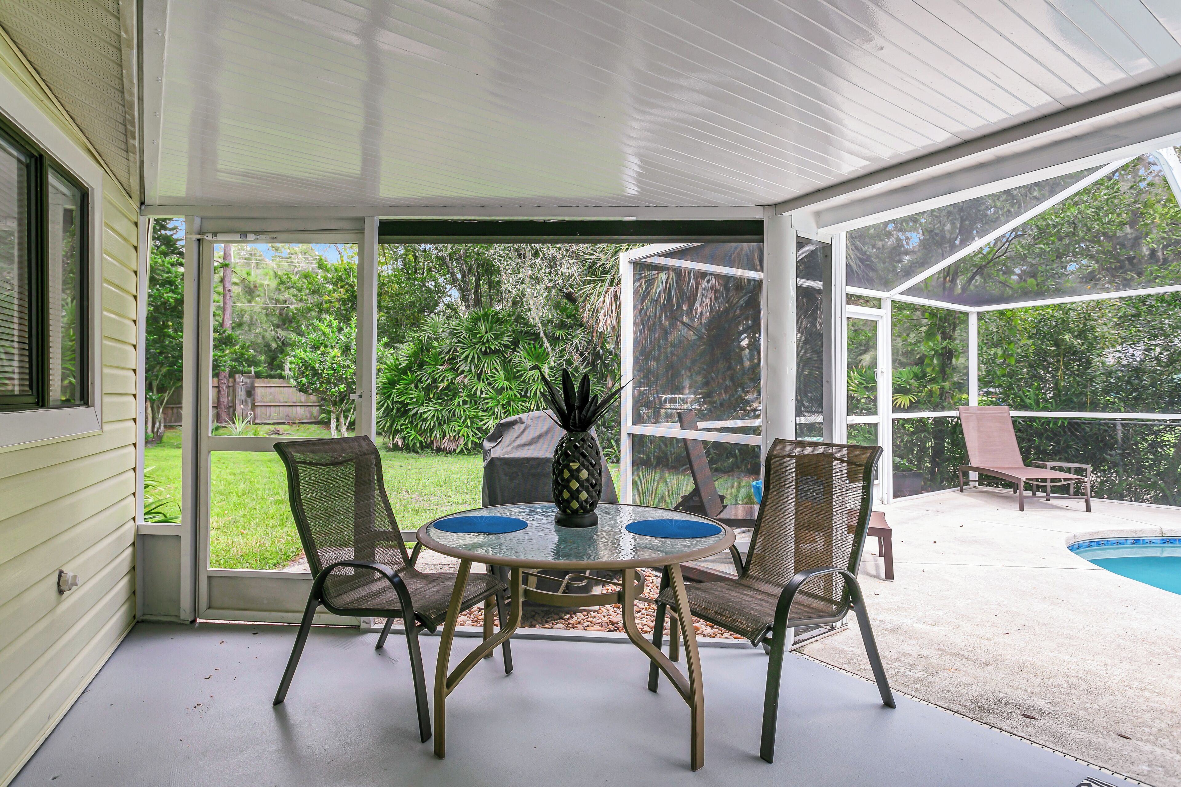 A covered patio with a glass door, a round table with chairs, and a pool in the background.