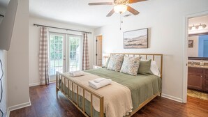 A bedroom with a wooden bed, a ceiling fan, a window with curtains, and a view of the kitchen.