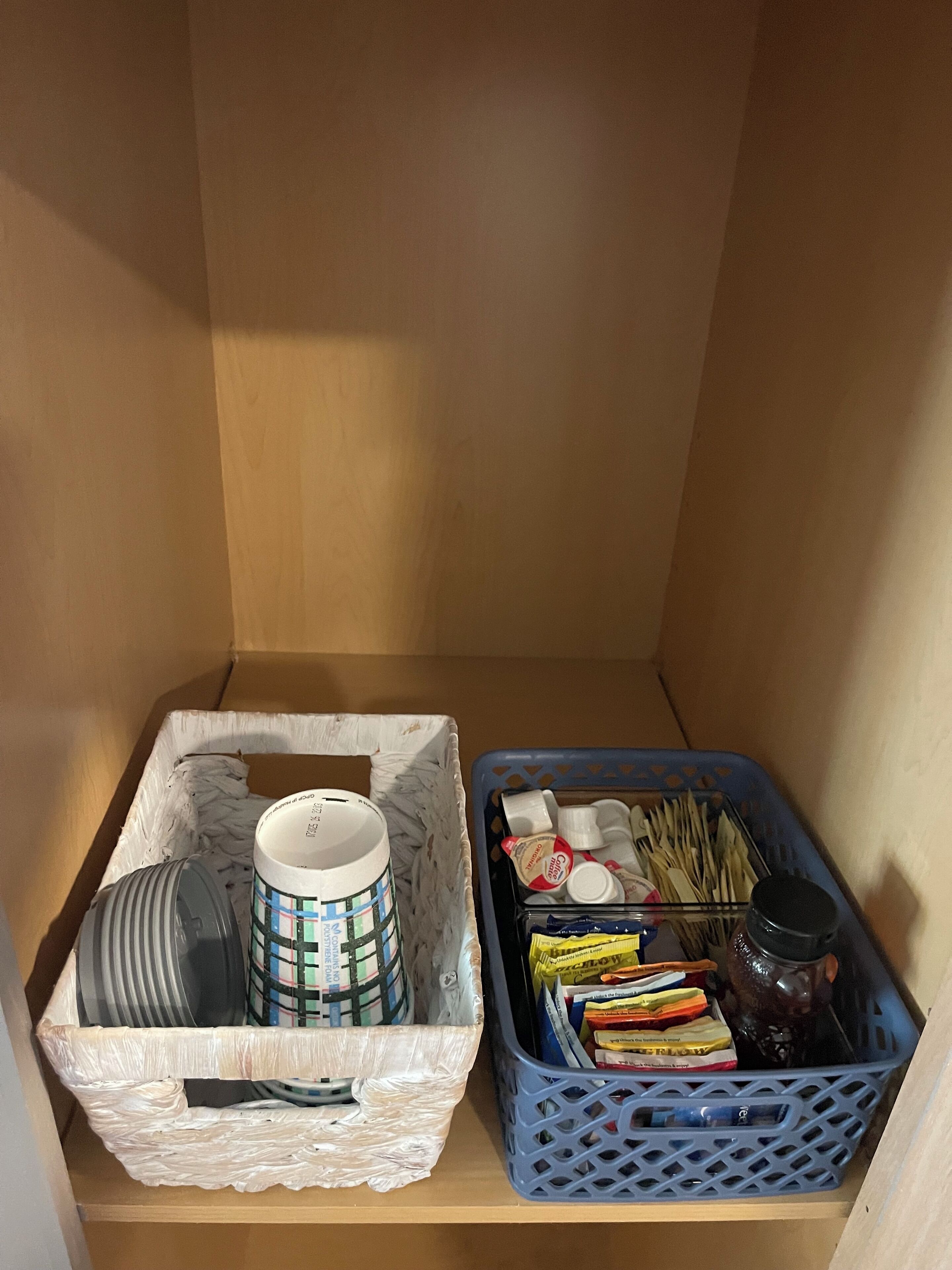 A storage area with a white basket containing a mug and a container, and a blue basket with food items and a thermos.