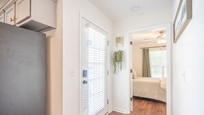 A hallway with a wooden floor, a door with a window, and a bed with a checkered bedspread in the background.