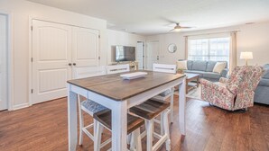 A spacious living area with a wooden dining table, white chairs, a floral armchair, a grey sofa, and a ceiling fan.