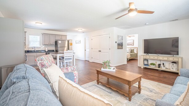 A spacious living room with a large sofa, a coffee table, and a television. There is a kitchen area with cabinets and a refrigerator in the background.
