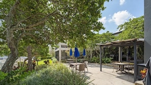 Une terrasse extérieure avec des tables et des chaises, abritée par une pergola et entourée de verdure et d’arbres.