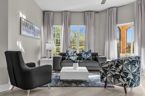 Una sala de estar moderna con un sillón negro, una mesa de centro blanca con un jarrón de flores, un sofá gris con cojines estampados y un sillón azul con motivos.