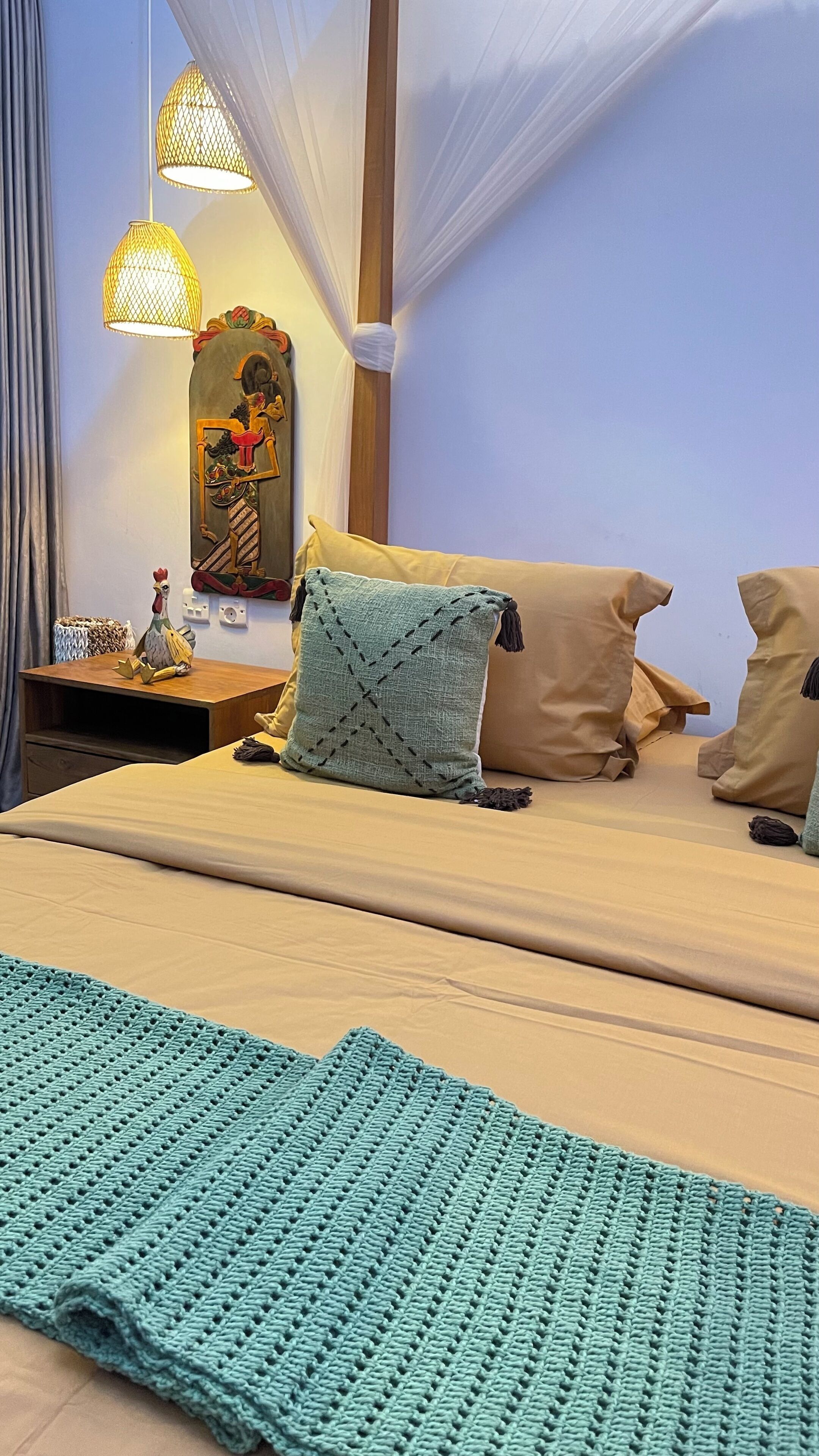 Un lit bien fait, recouvert d’un plaid vert en tricot, une table de chevet en bois ornée d’un coq décoratif et une lampe fixée au mur.