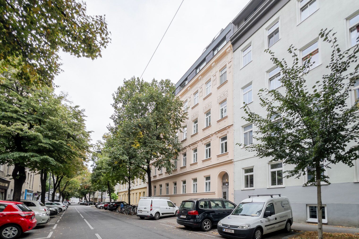 Una calle bordeada de coches estacionados y árboles, con un edificio de varios pisos que presenta un estilo arquitectónico clásico al fondo.