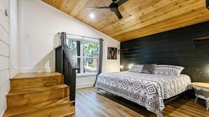 Un dormitorio con escalera de madera, cama con ropa de cama estampada, ventilador de techo y ventana con cortinas.