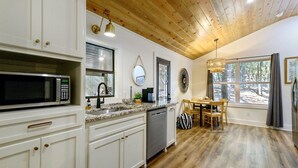 Una cocina moderna con armarios blancos, un microondas de acero inoxidable y un lavavajillas. Hay un comedor con mesa y sillas de madera, y un ventanal con vistas a árboles.