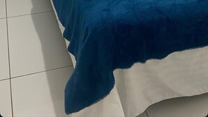 A bed with white bedding and a blue blanket, a small black stool, and a white wall with a blue panel. - Pousada Santo Mel (São Tomé)