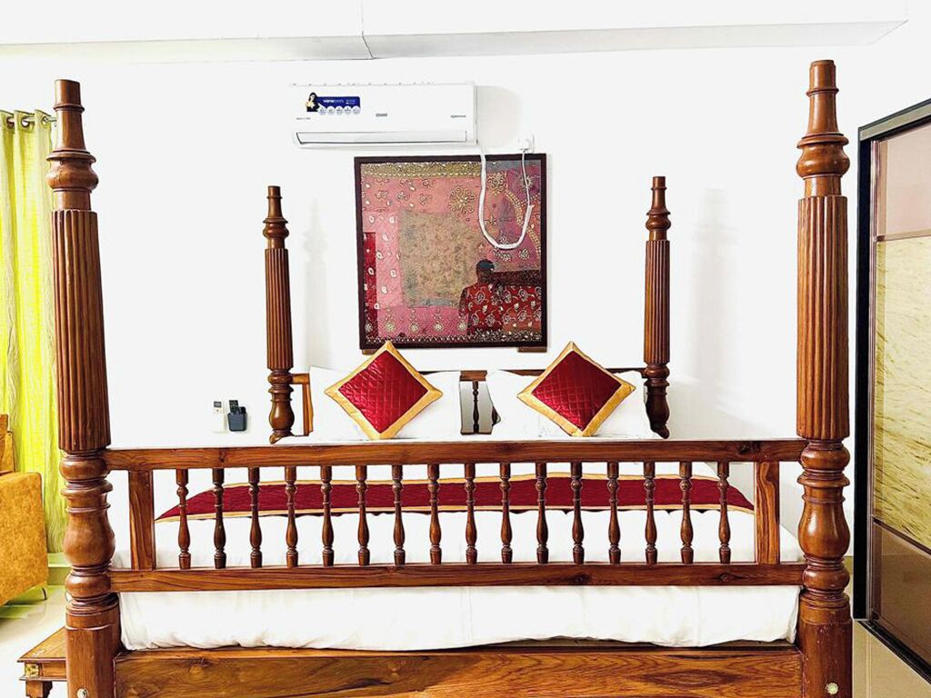 A wooden four-poster bed with a red and white bedspread, a framed picture on the wall, and a small air conditioning unit above it.