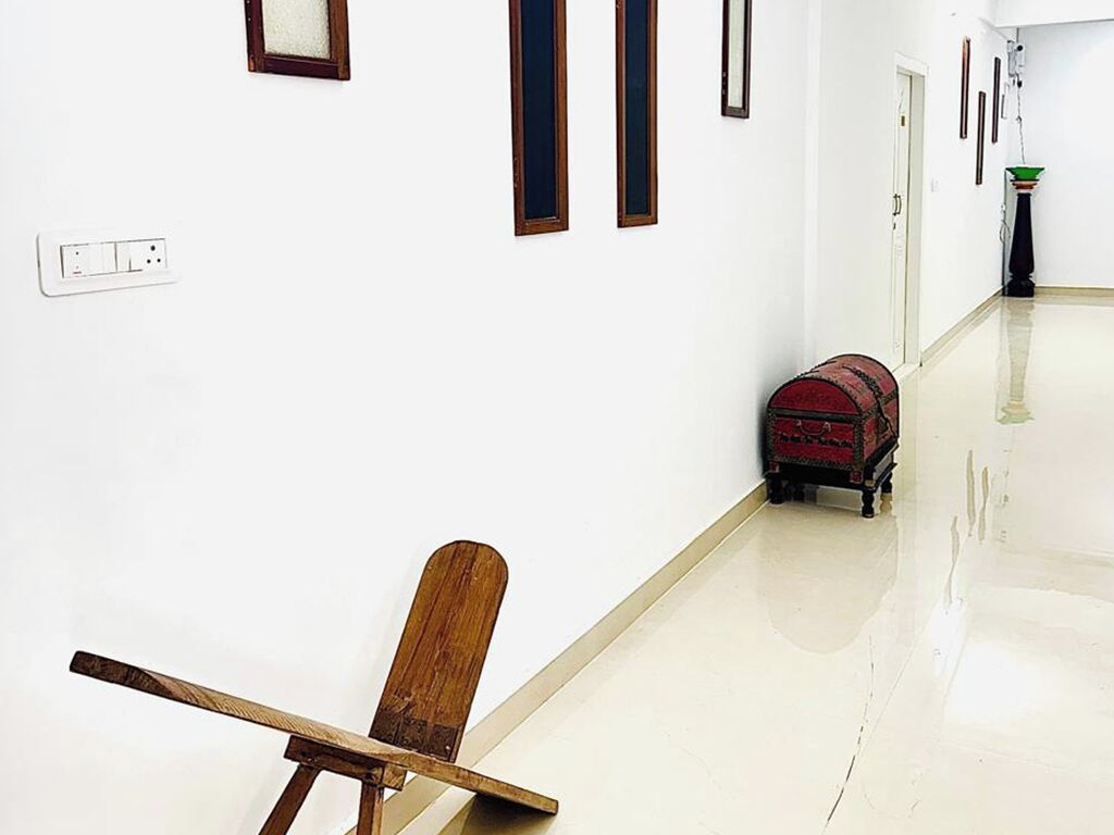 A hallway with a wooden chair, a red suitcase, and a white wall with a power outlet.