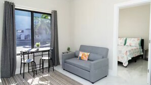 Una habitación moderna con un sofá gris, una pequeña mesa con sillas, una cama con ropa de cama floral y vistas a palmeras a través de amplios ventanales.