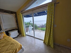Ein Schlafzimmer mit Bett, einem Fenster mit Vorhängen, einer Glastür, die zu einem Außenbereich mit Tisch und Stühlen führt, und Blick aufs Meer.