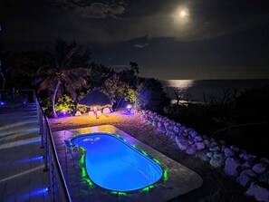 Ein nächtliches Bild mit einem Pool, einem Gebäude mit einem Reetdach und Blick auf das Meer unter dem vollen Mond.
